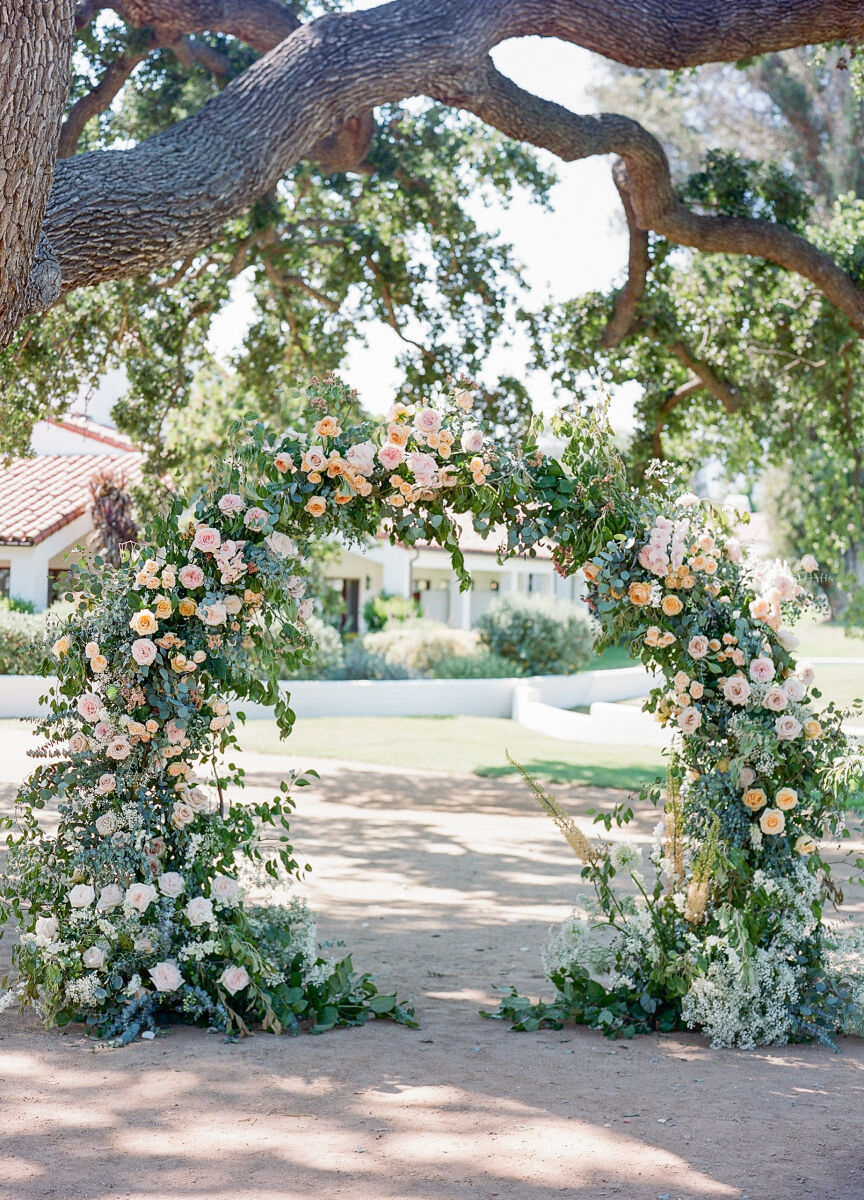 A lush arch of pink and peach blooms at an outdoor wedding ceremony.