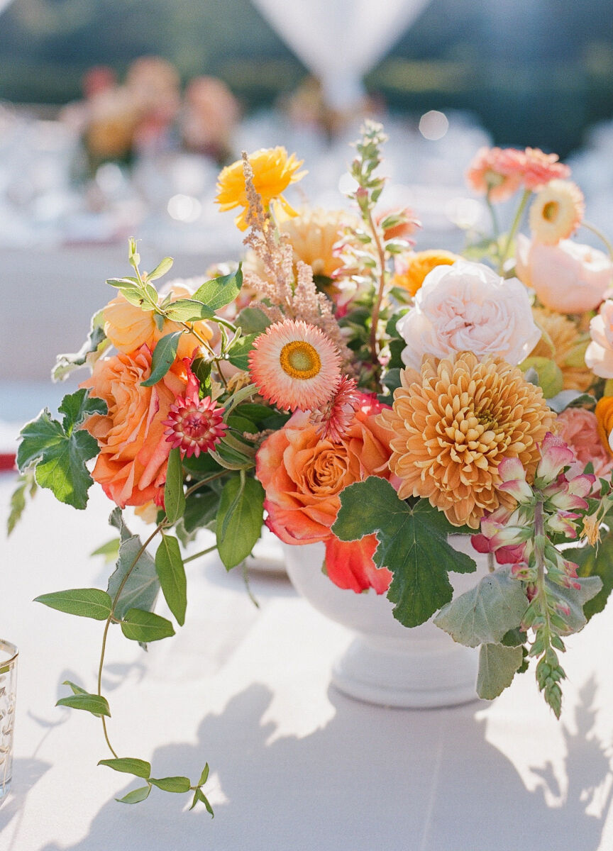 A bright wedding centerpiece of peach, orange, and yellow flowers with trailing greenery in a white compote.