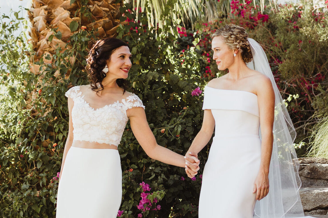 Pink Wedding: Two brides smiling and holding hands, while walking in a desert garden on their wedding day in Palm Springs, California.