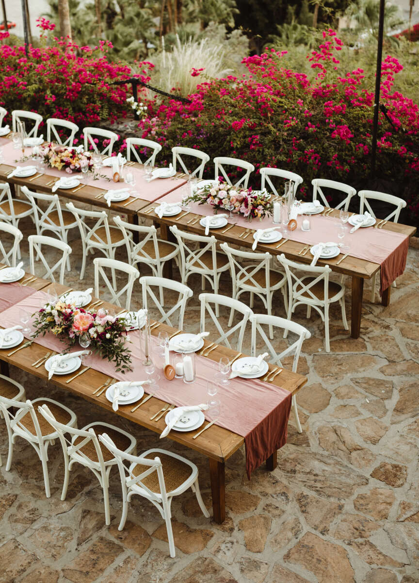 Pink Wedding: Reception tables set with blush pink table runners and gold flatware on a garden terrance surrounded with bright pink flowered bushes.