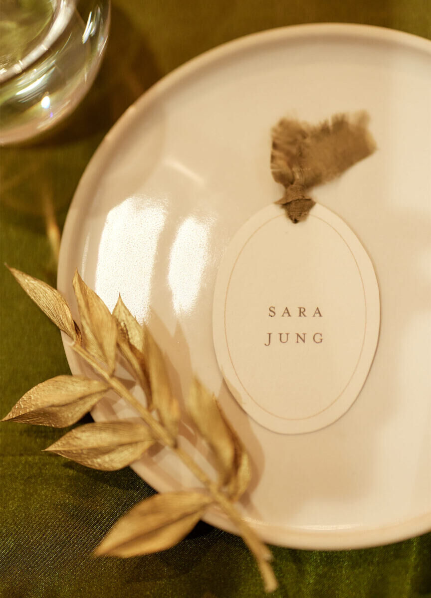 An oval place card with silk ribbon detail, sitting on a dinner plate by some gilded greenery at a restaurant wedding reception.