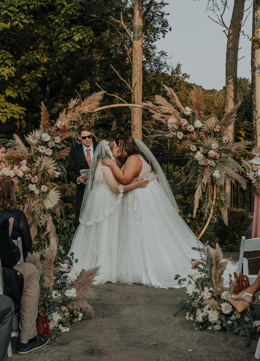 Romantic Wedding Venues: Two brides kissing under a floral arch outdoors at Grove at Briar Barn Inn.