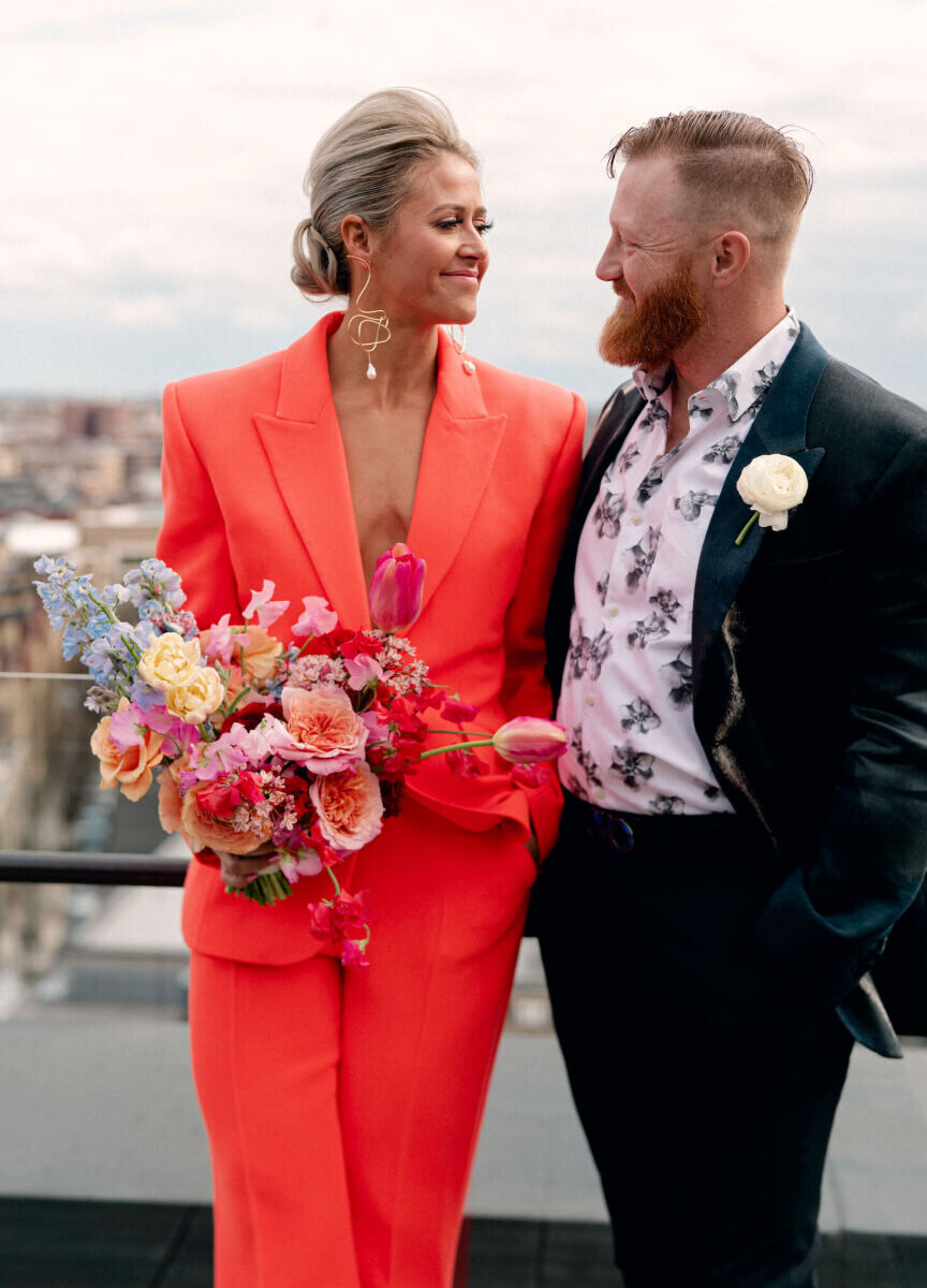 A couple lock eyes at their surprise wedding on a rooftop in DC—she in an orange suit holding a colorful bouquet, and he in a chic blazer adorned by a modern boutonniere.