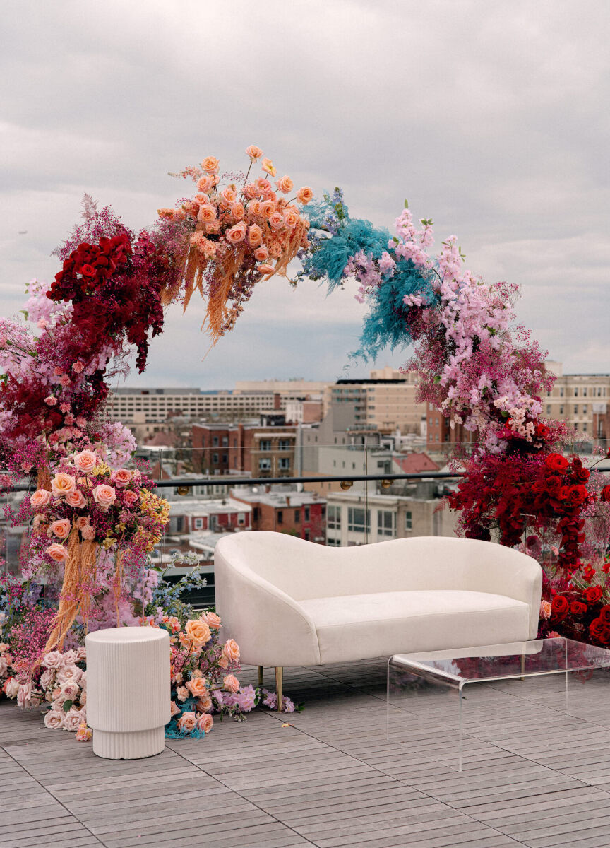 An arch made of clusters of colorful flowers, surrounds a white and clear lounge area on a rooftop, that then became the background of a surprise wedding ceremony.