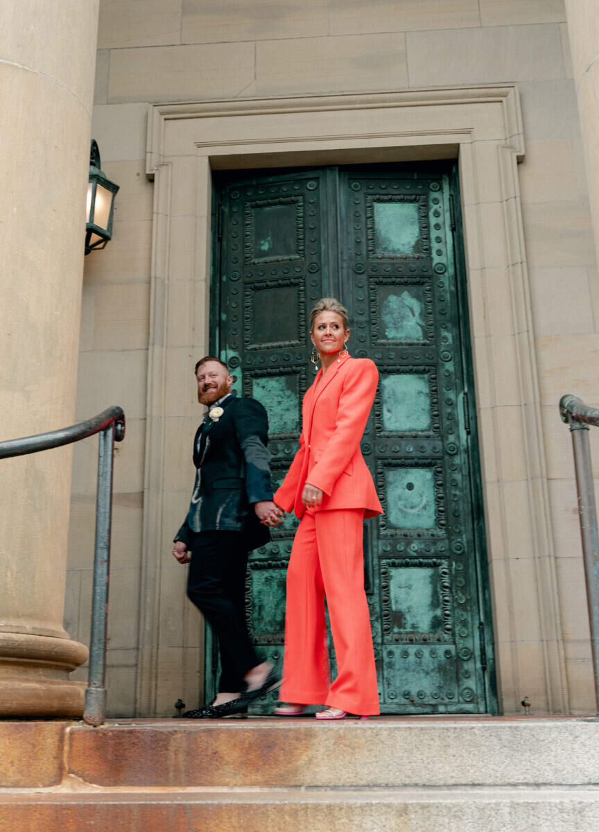 A bride in an orange suit, led by her groom in a statement blazer, walk in front of a teal door, at the front entrance of the venue for their surprise wedding—a hotel in Washington D.C.