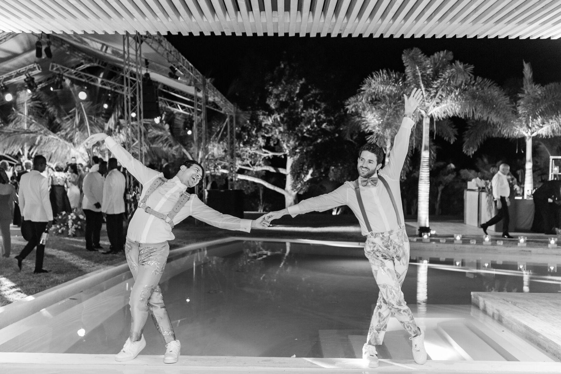 Tropical Wedding: Two grooms holding hands while standing on the edge of a pool during their wedding reception.