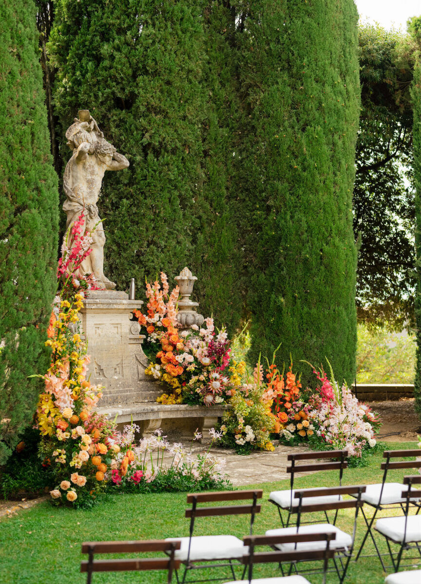 A Tuscan wedding ceremony setup with a statue as the background and colorful flowers surrounding it.