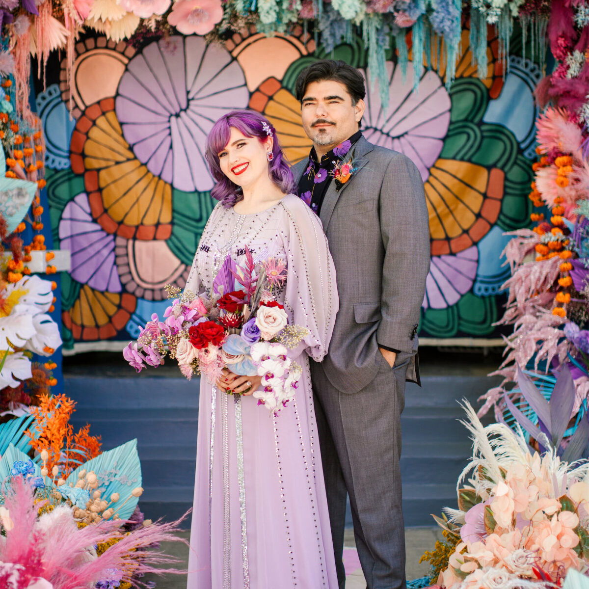 A bride and groom pause for a portrait at their vibrant outdoor wedding, which incorporated a rainbow floral installation and painted backdrop at their ceremony.