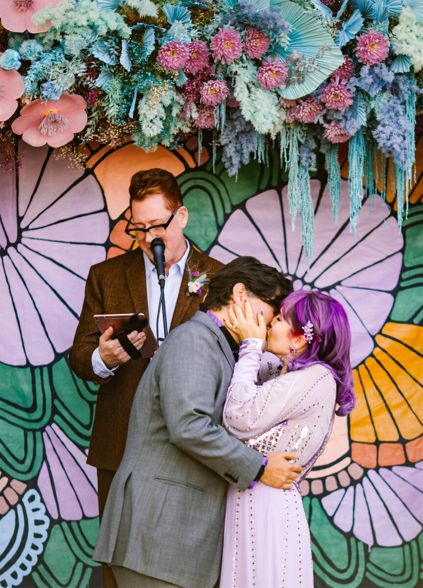 A groom and bride embrace in front of a painted backdrop at their vibrant outdoor wedding.