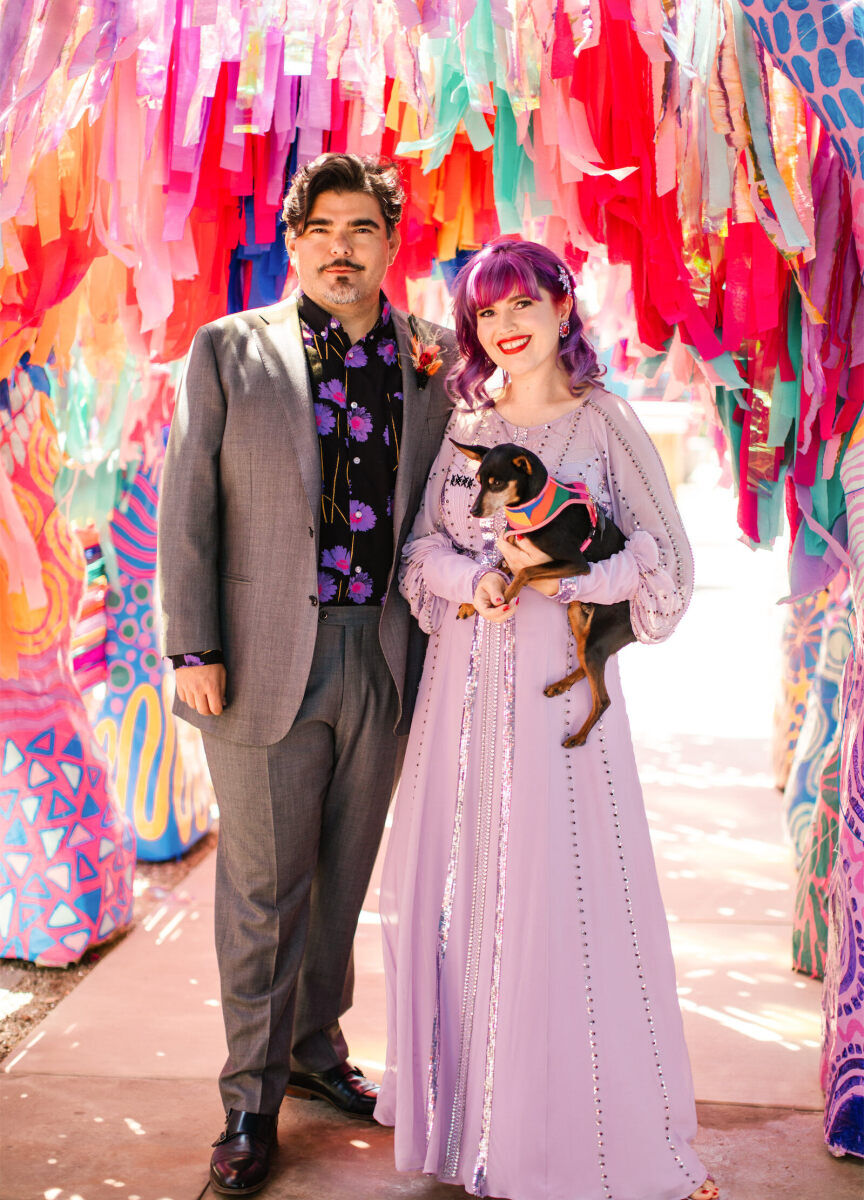 A couple and their miniature pinscher pose for a portrait in the mixed-media art installation tunnel that was a focal point of their vibrant, outdoor wedding.