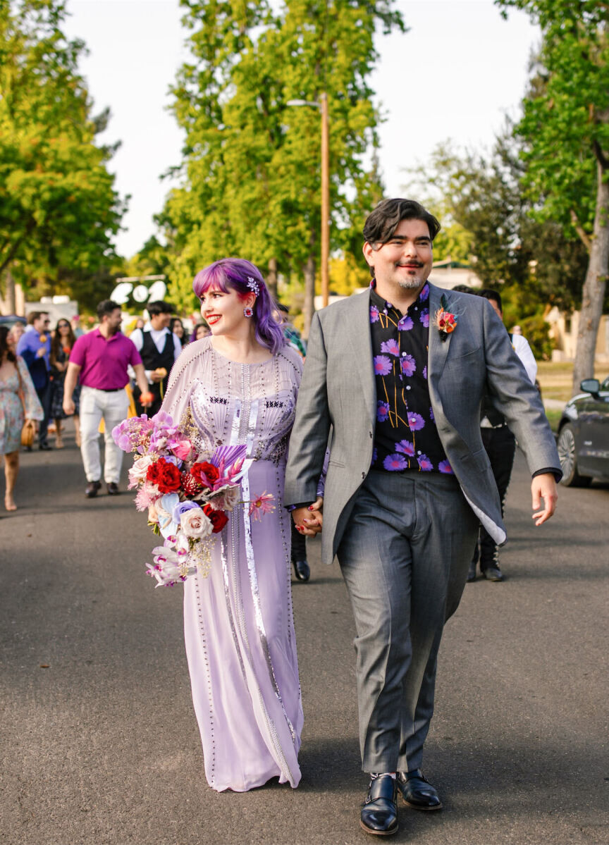 A bride, wearing a lavender dress, and a groom, in a grey suit and purple shirt, smile as they parade through the streets of their neighborhood following their vibrant, outdoor wedding ceremony.