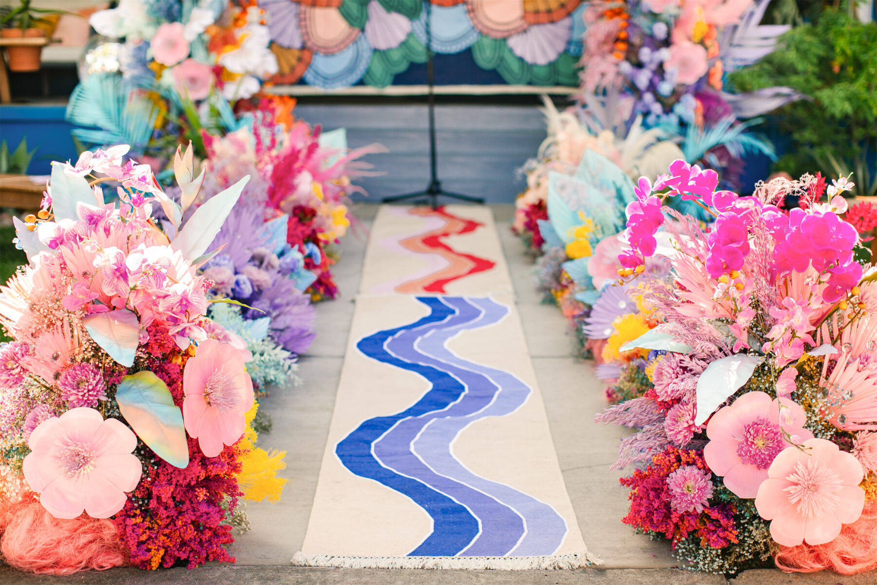 A rainbow of flowers (a mix of real, dried, and faux) line a painted aisle leading towards the front porch of a couple's California home where their vibrant outdoor wedding took place.