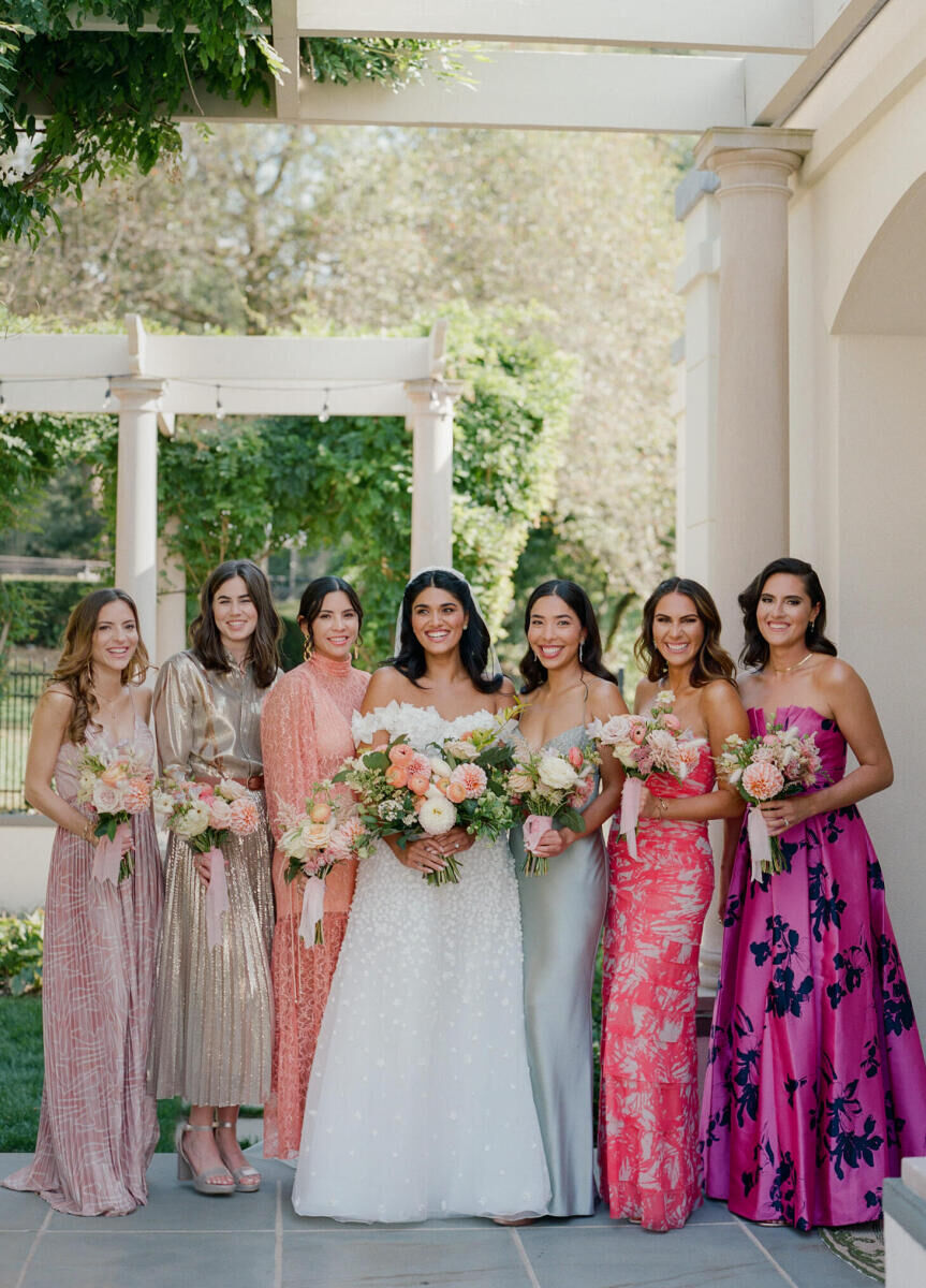 Bridesmaids wearing mismatched dresses in shades of pink and metallics surround the bride, and they all hold vibrant bouquets and smile.