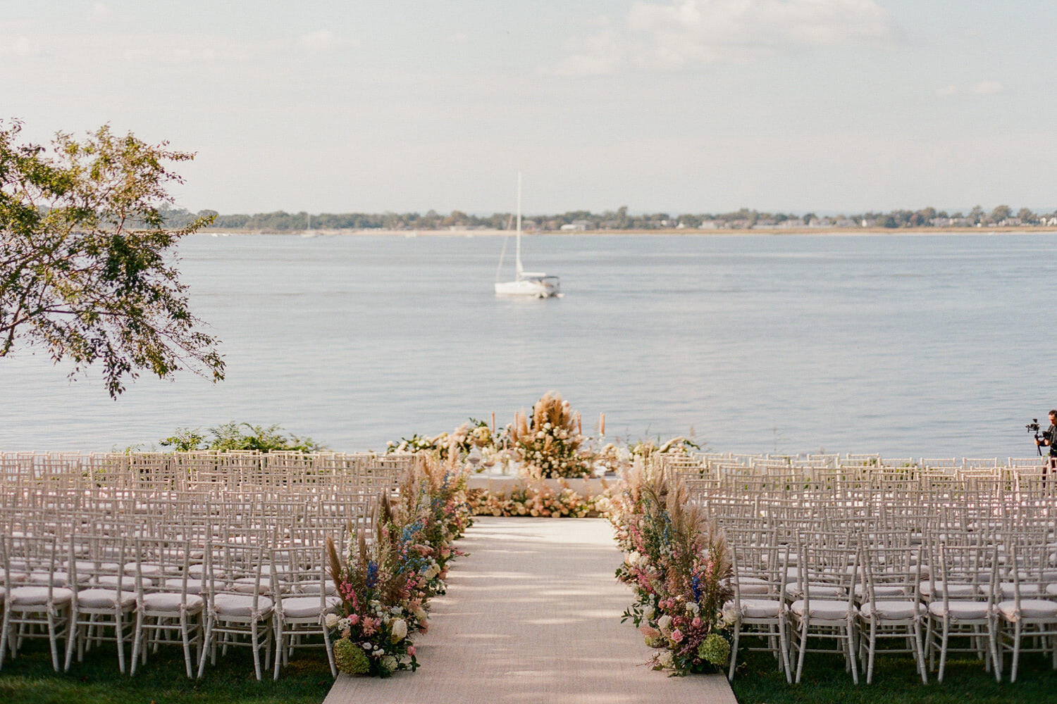 A waterfront wedding ceremony overlooking Oyster Bay in Long Island is set with white Chiavari chairs and an aisle lined with pastel flowers.