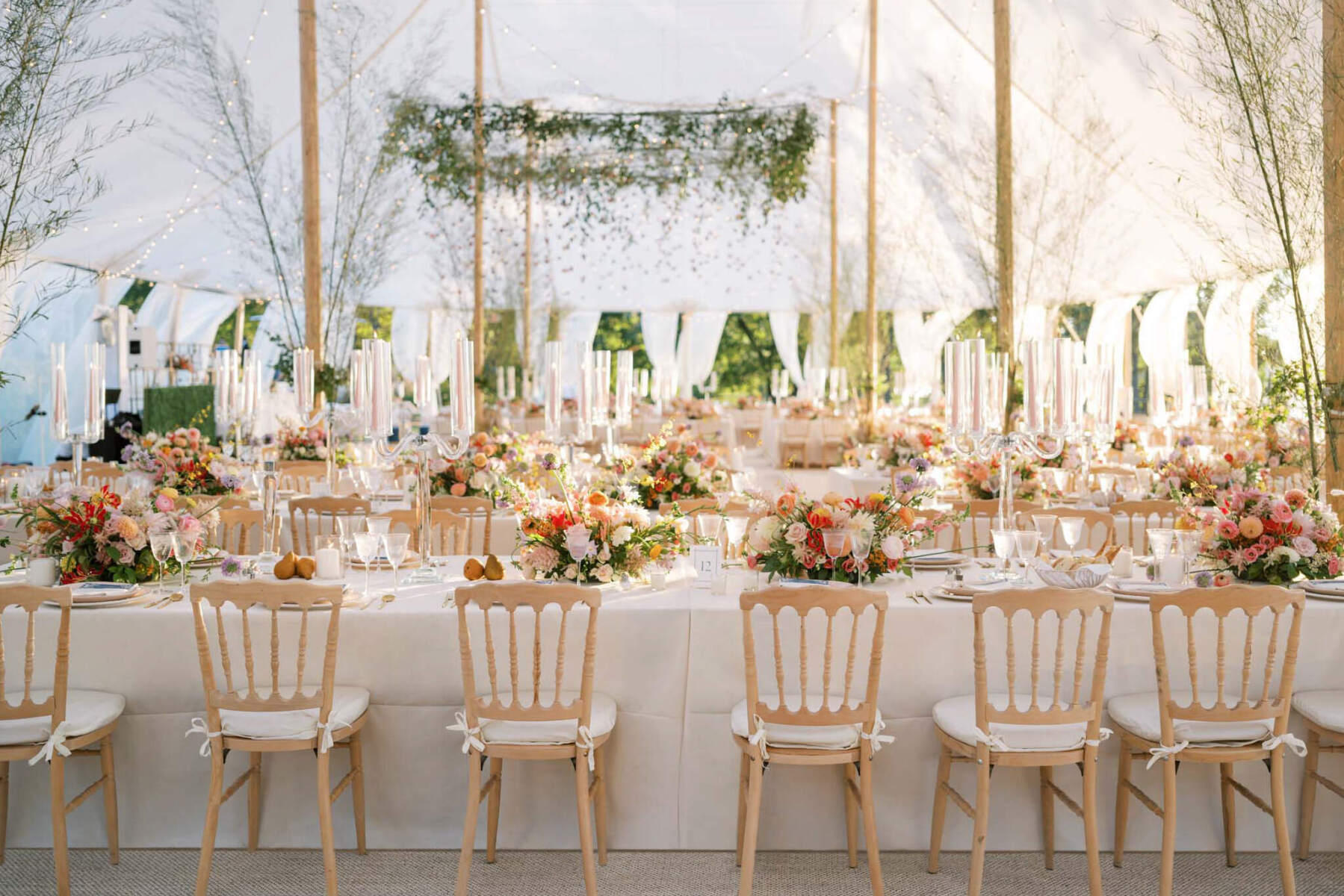 The tented reception of a waterfront wedding, decorated with a canopy of greenery and folded paper cranes over the dance floor and centerpieces made with a variety of warm-hued flowers and fruit, paired with elegant pink taper candles.
