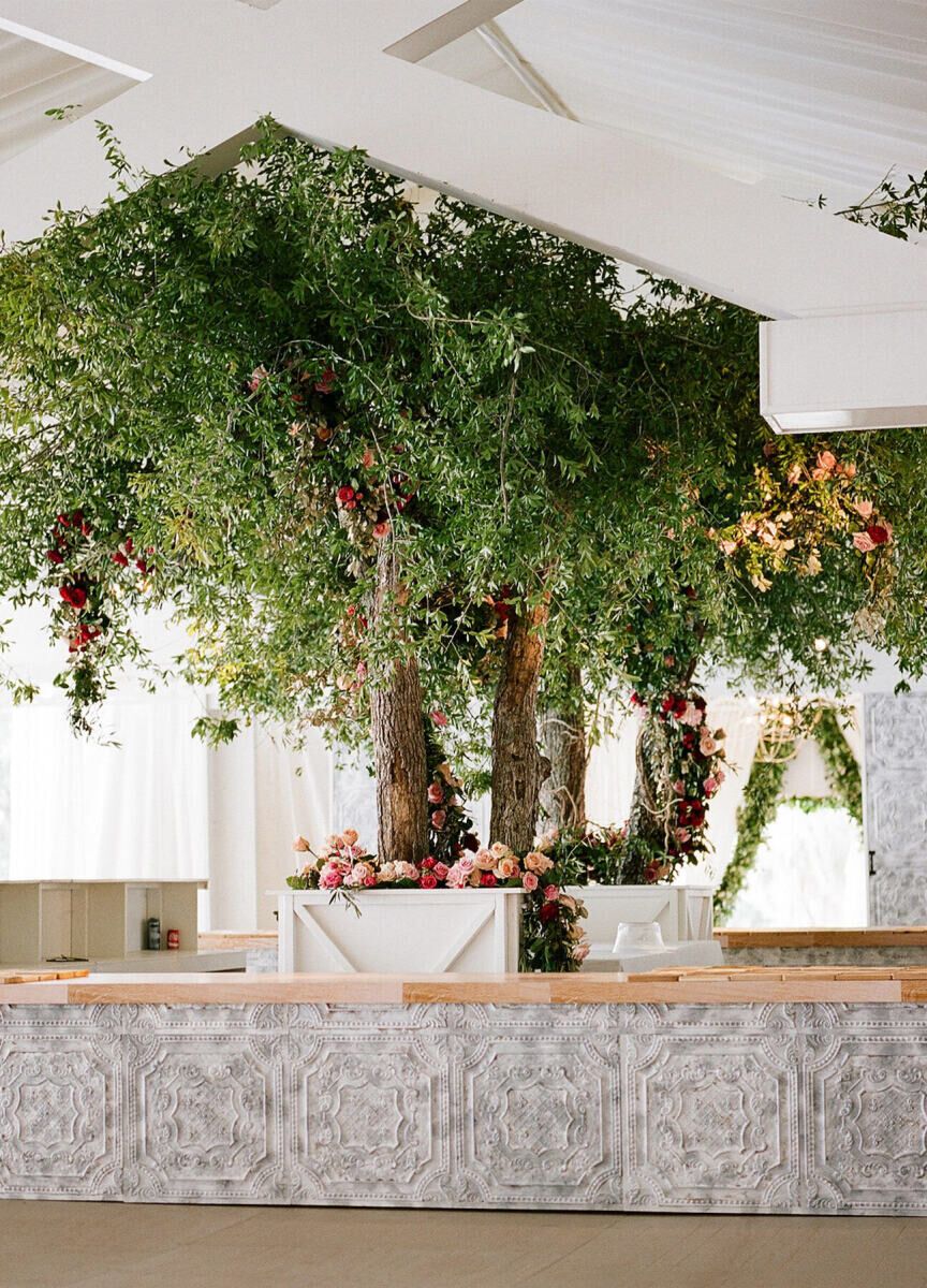 Wedding budget: Wedding reception bar featuring detailed marble grey paneling and large tree installation in the center surrounded by pink and red florals 