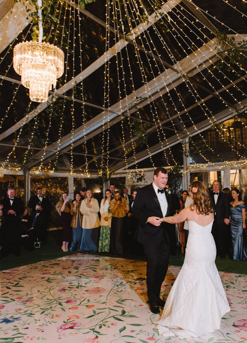 A bride and groom perform their first dance on a floral-patterned dance floor in a clear tent.
