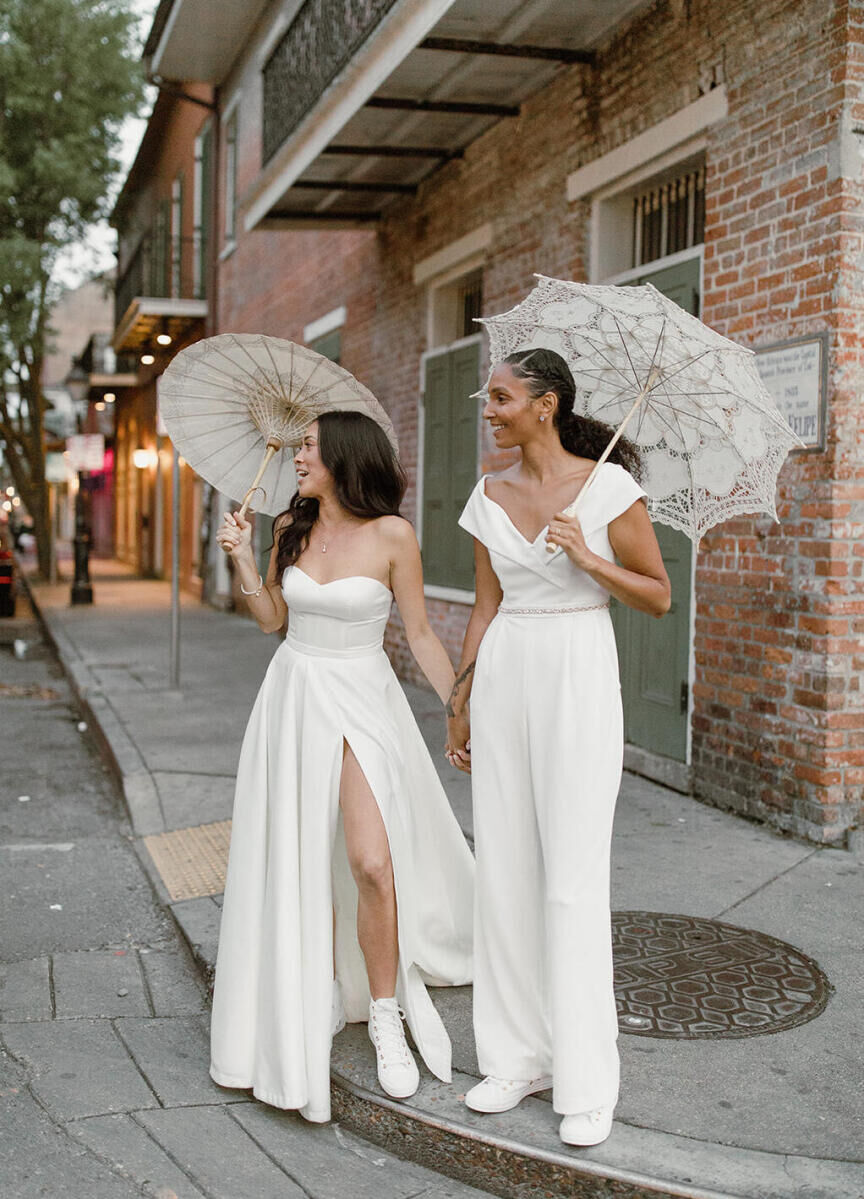 Wedding Dress Shopping: Two brides, one in a high-slit gown and the other in a white jumpsuit, holding hands while holding parasols in New Orleans.