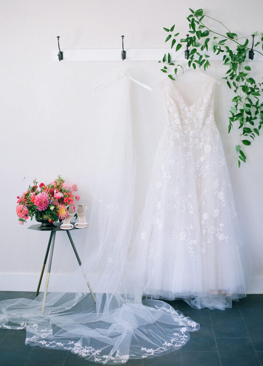 Wedding Etiquette Budget: A pair of wedding shoes on a table with a colorful floral arrangement, next to a veil and wedding dress hung up nearby.