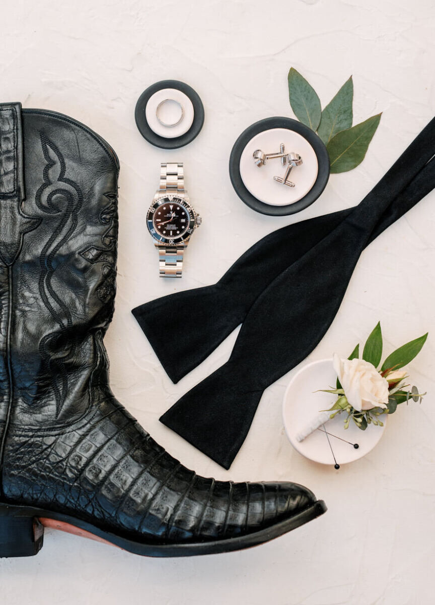 Wedding Etiquette Budget: A flat-lay of a black cowboy boot, wedding ring, watch, undone bow tie, cufflinks, and boutonniere.