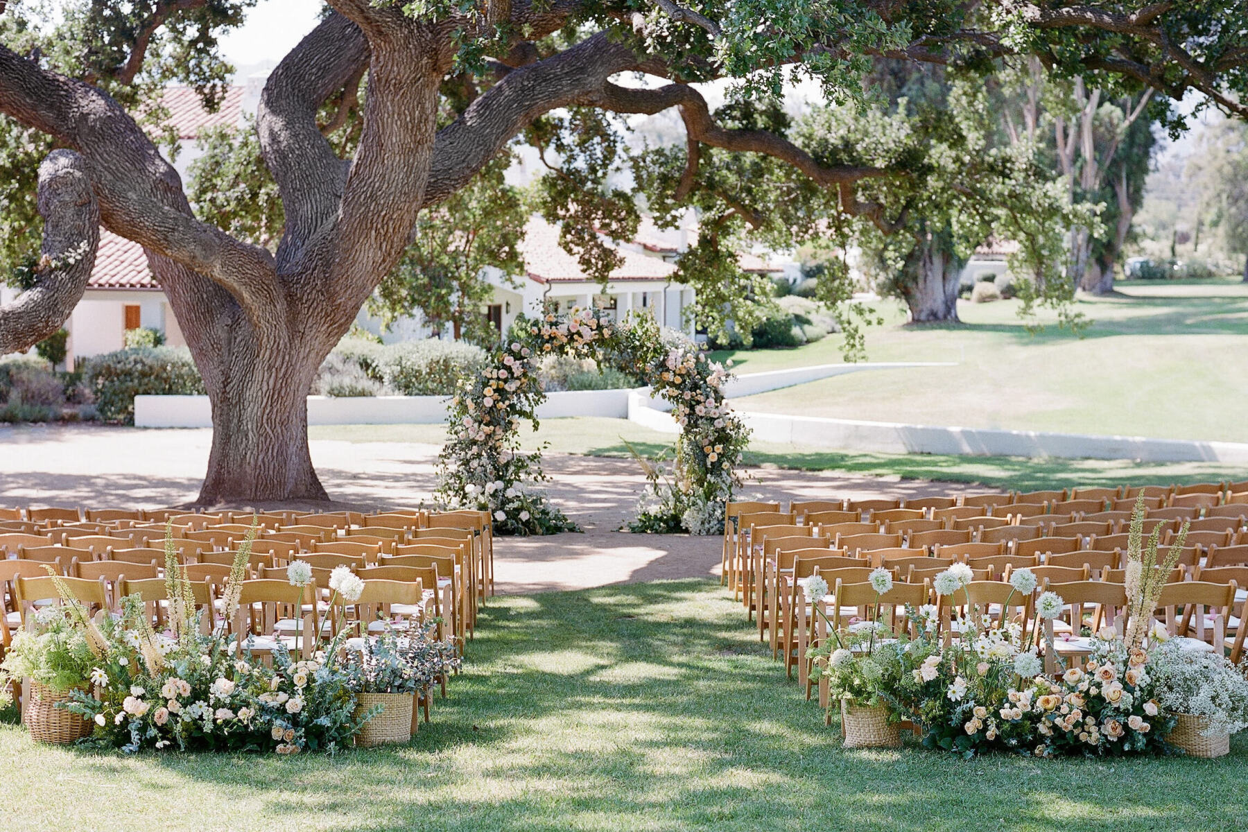 Wedding Etiquette: Wedding reception setup under a large oak tree, with an aisle lined with spring flowers and greenery, leading to a wedding arch made of the same flowers.
