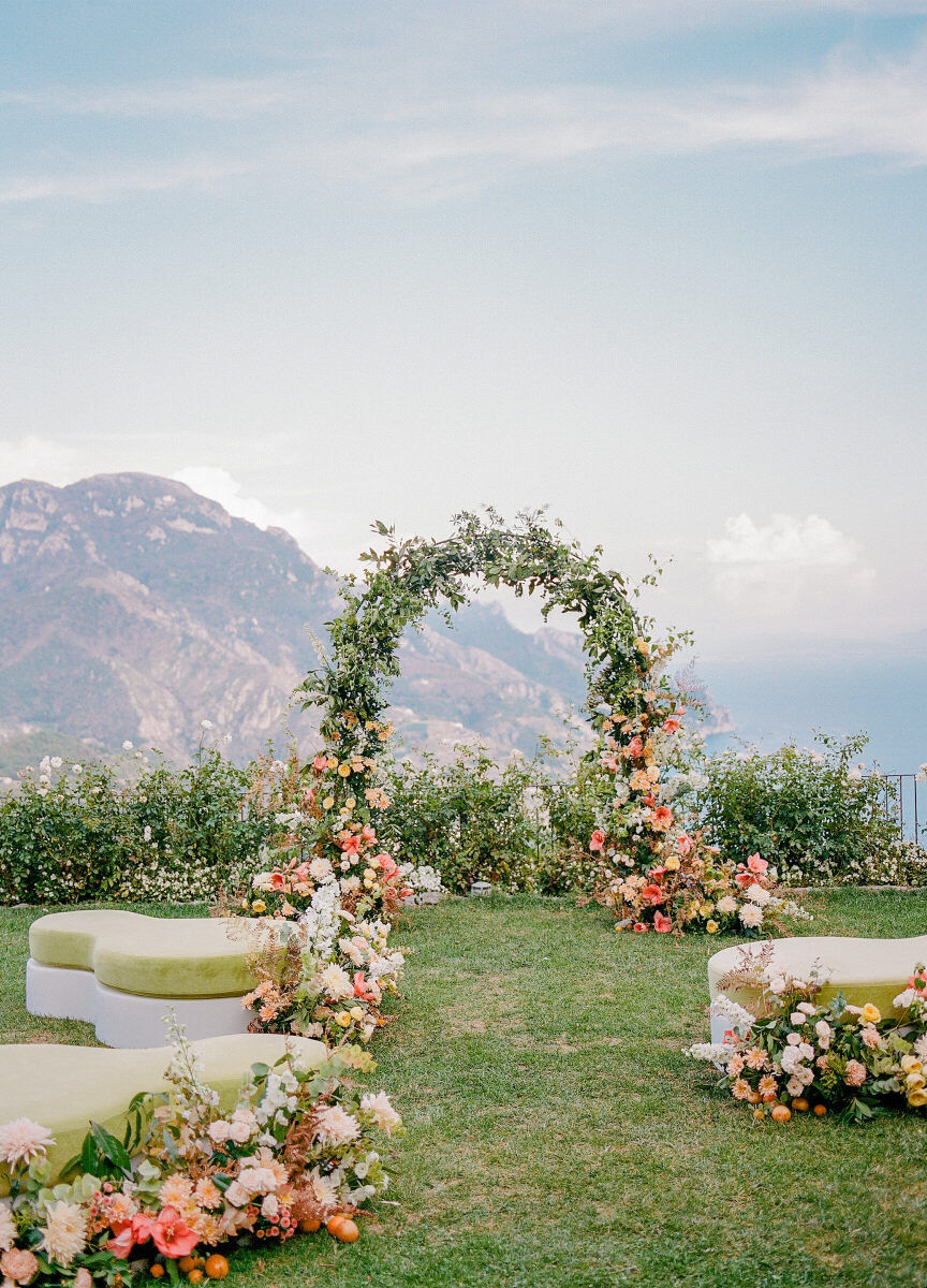 An outdoor ceremony setup with a floral arch overlooking the mountains and sea, with cushioned green benches leading up to the altar.
