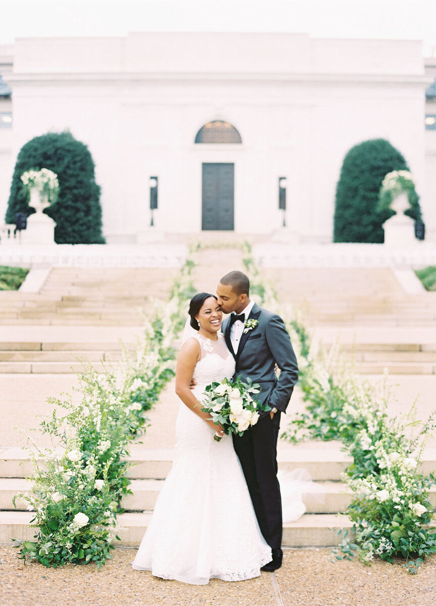 A groom kissing a bride at the bottom of an outdoor staircase with green and white arrangements leading up to an event space in DC.