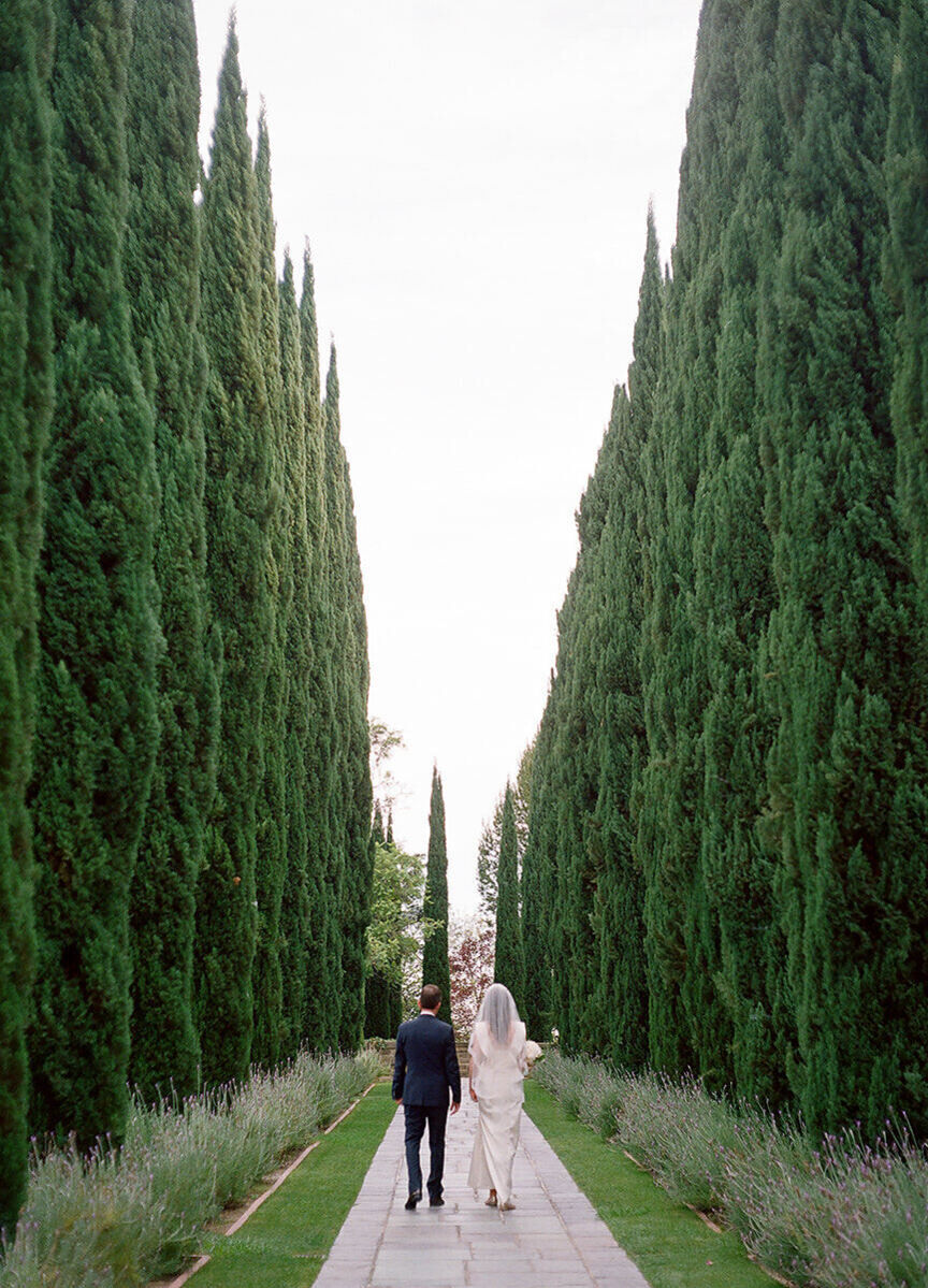 Wedding Photography Ideas: A wedding couple walking away from the camera, along a tall hedge plant-lined walkway.