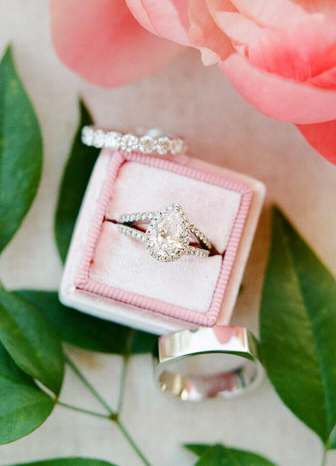 Wedding Ring Design: A pear cut engagement ring sitting in a white and pink box surrounded by two wedding bands against a floral backdrop.