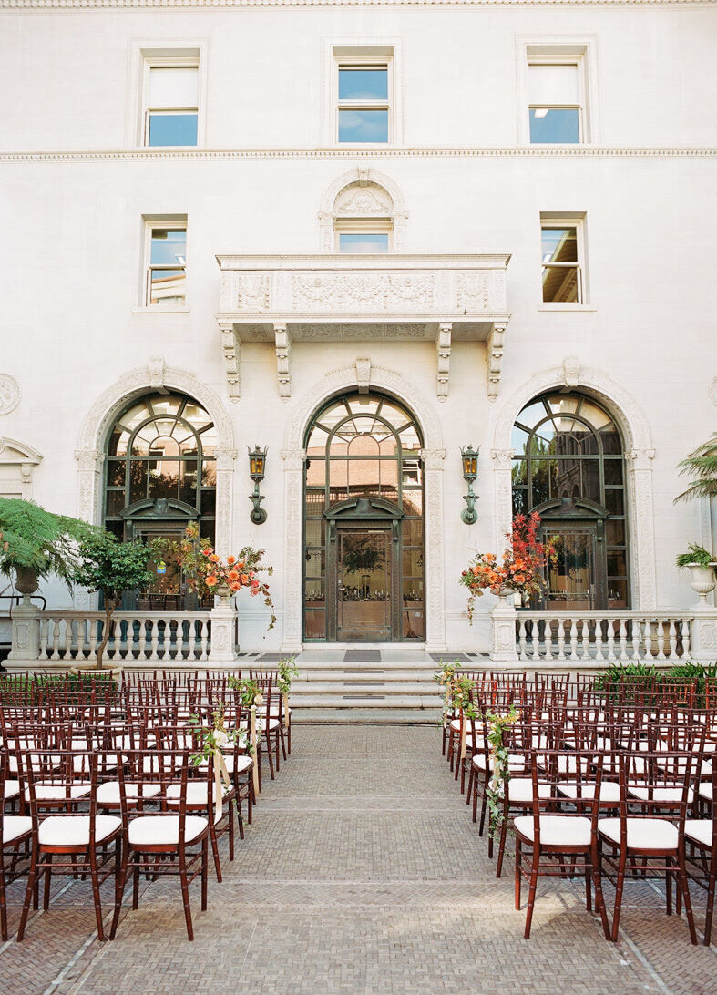 Wedding Tips: An outdoor ceremony set-up in front of a white mansion. There are wooden chairs set up facing the steps to the mansion.
