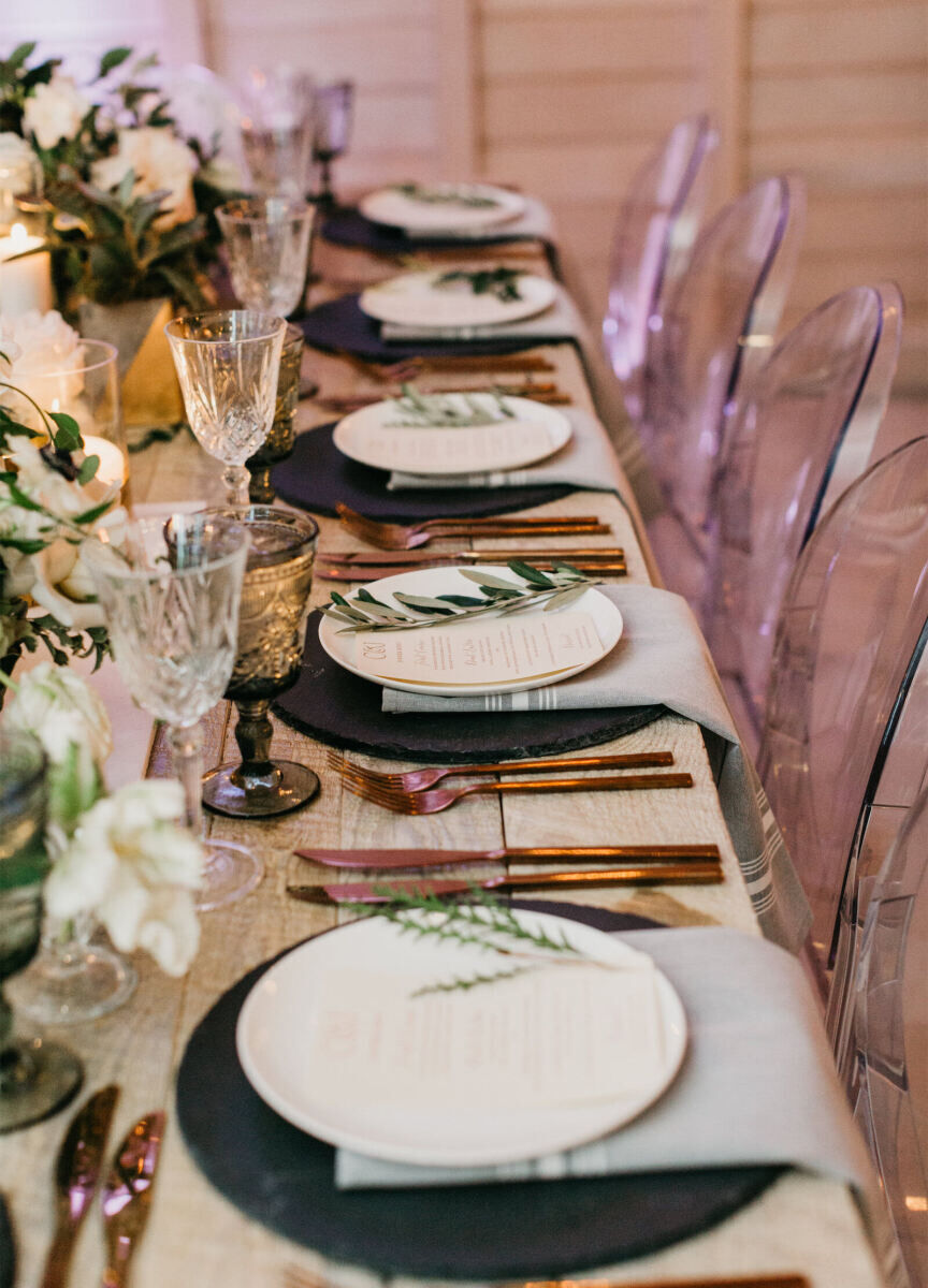 Wedding Tips: Photo of a ceremony dinner table set up. There are five white plates, each adorned with a menu and piece of greenery. There are embellished glasses and clear chairs surrounding the plates.