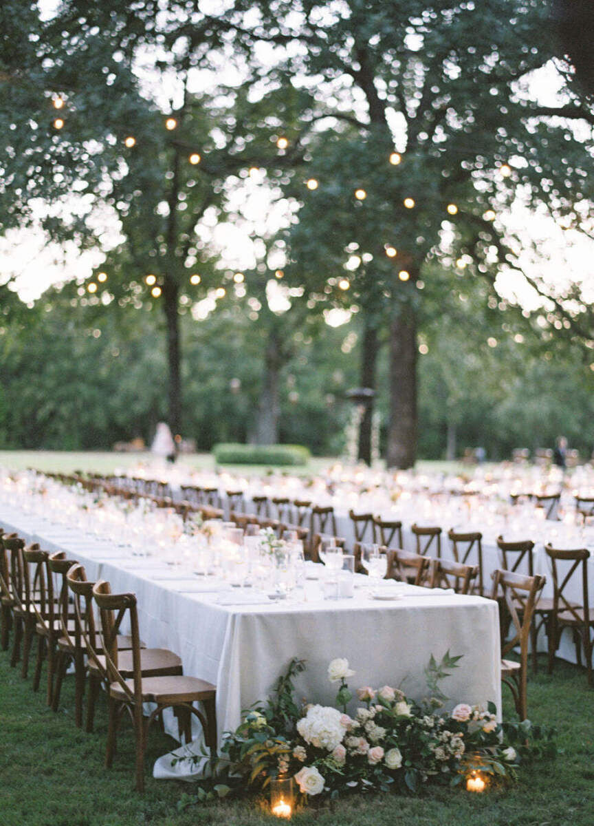Wedding Tips: Long, large tables with white tablecloths and wooden chairs set up for an outdoor wedding reception under string lights.