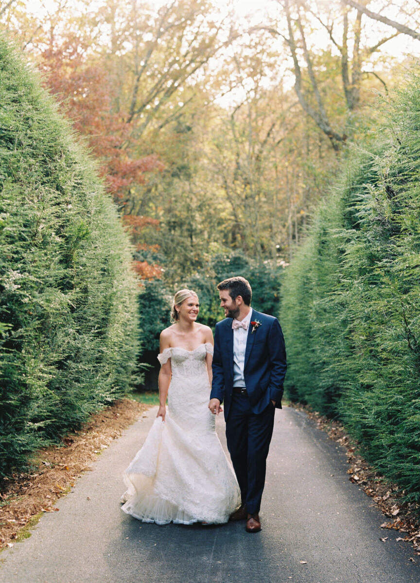 Wedding Tips: A bride and groom smiling at each other while holding hands and walking down a tree-lined path outdoors together.