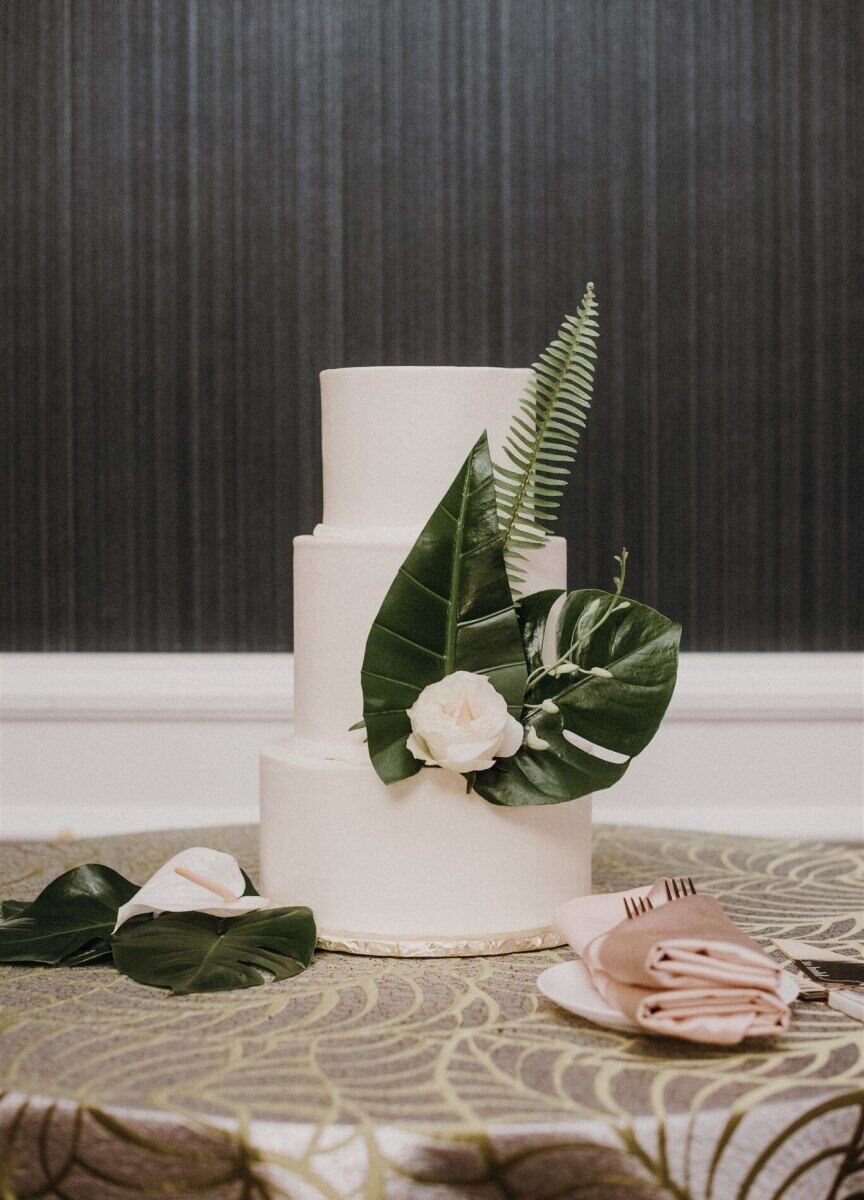 Wedding Website Examples: A three-tiered white wedding cake with tropical leaves and a white flower as accents.