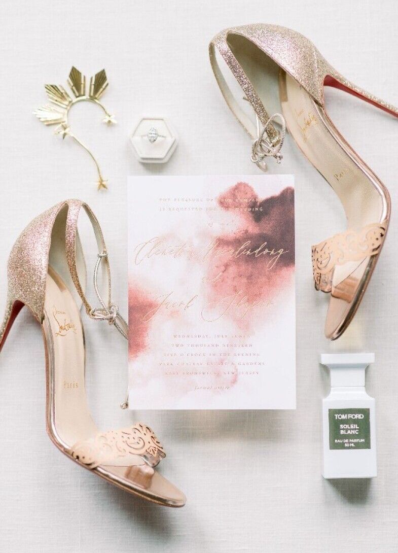 Wedding Website Examples: A flat lay of a red and pink invitation, gold wedding shoes, perfume, an engagement ring, and more.
