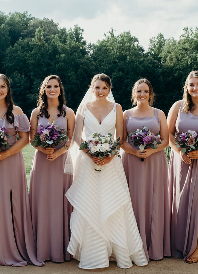 Wedding Website Examples: A bride posing outdoors with her four bridesmaids, who are all wearing lilac-colored dresses.