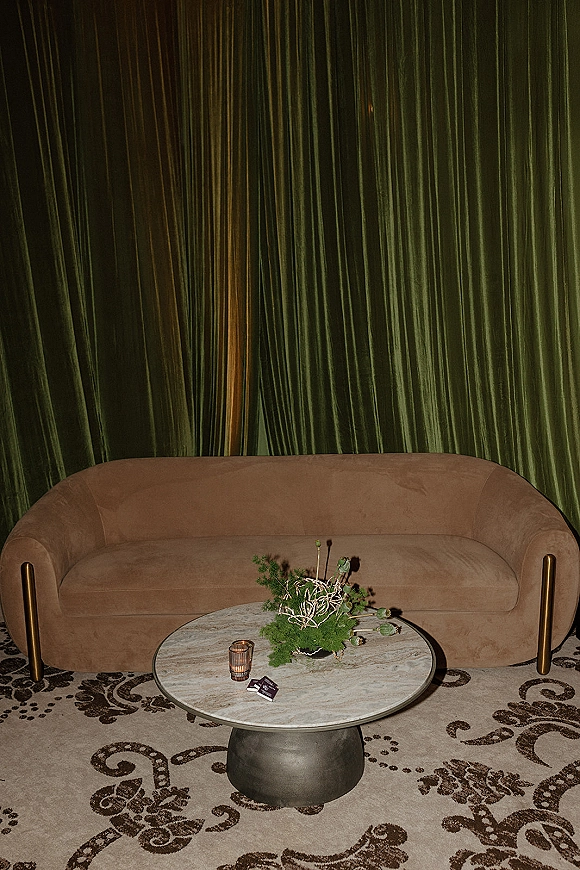 Wedding lounge area with a velvet sofa and marble coffee table, floral centerpiece and votive candle, set against green velvet curtains