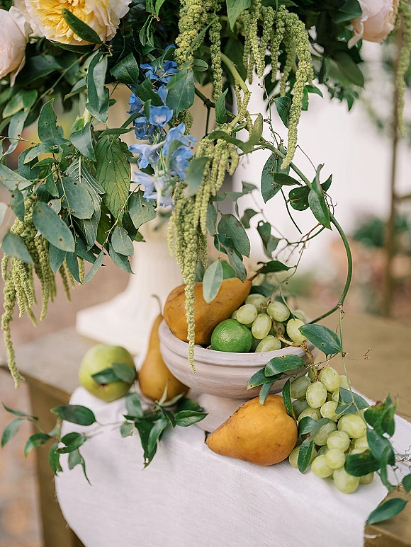 Wedding tablescape with a fruit wedding centerpiece of green grapes, pears, limes, and yellow roses in a ceramic bowl on white linen on a wood table