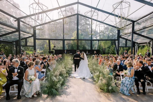 Ceremony moment as bride and groom walk down the aisle in a clear top tent, veil flowing, guests cheering beside white florals and greenery