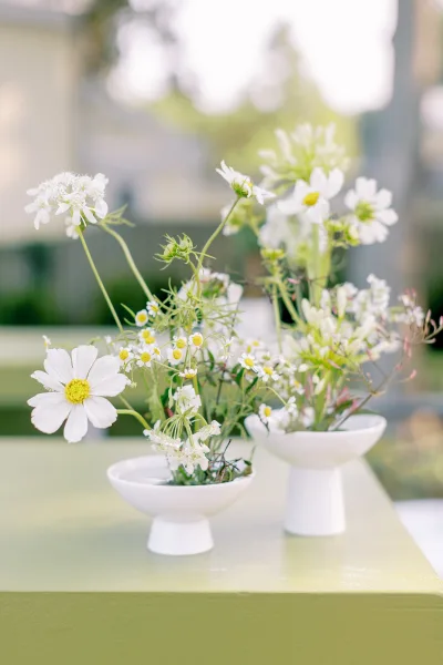 Wedding centerpiece with wildflower wedding centerpiece daisies in white pedestal bowls and bud vases, greenery stems, blurred garden backdrop