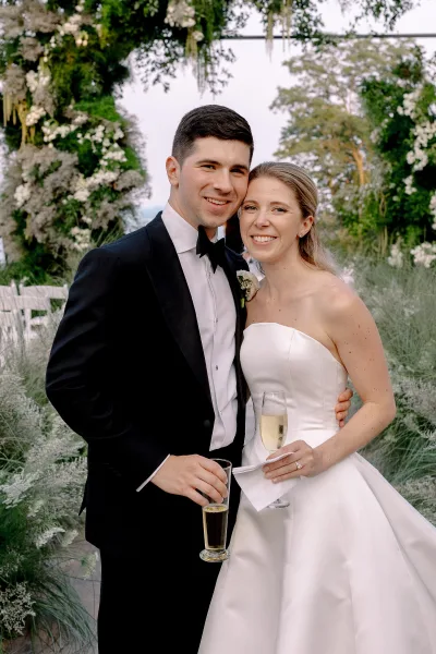 Couple portrait of a smiling bride and groom holding champagne flutes, with strapless dress and black tuxedo by a white floral arch outdoors