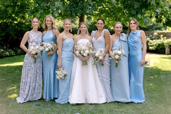 Bridesmaid group photo with bride in veil and white gown, bridesmaids in blue dresses holding white bouquets in a sunny garden lawn setting