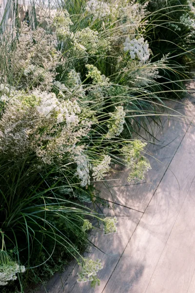 Ceremony aisle florals with grounded white flowers, ornamental grasses, and greenery lining a wooden aisle in an outdoor garden setting