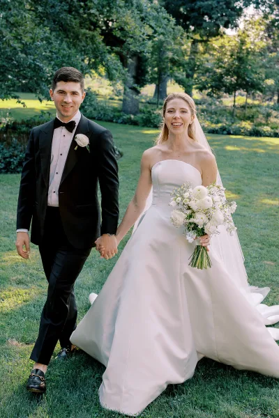 Couple portrait of bride and groom holding hands, bride in veil with bouquet beside groom in tuxedo on a green garden lawn
