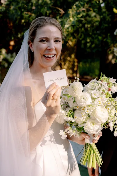 Bride portrait of a smiling bride holding bouquet and escort card, veil over shoulders in sunlit garden greenery with trees