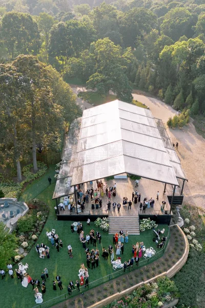 Wedding reception under a clear roof tent with cocktail tables and white chairs on a lawn beside hydrangea bushes and forest trees