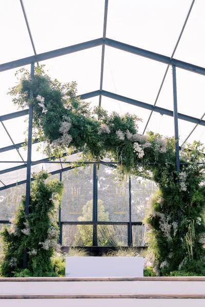 Wedding ceremony arch with a greenery wedding arch and white flowers, framed by hanging chandeliers on a stage inside a glass greenhouse