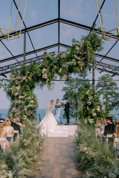 Wedding ceremony setup with a floral ceremony arch and greenery garland under a clear tent, white chairs facing an ocean view backdrop