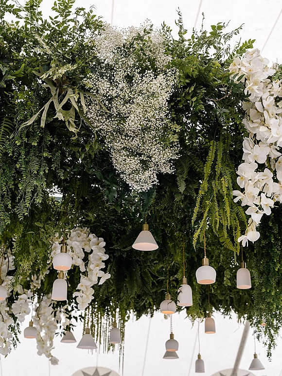 Hanging floral installation with wedding ceiling greenery, baby’s breath and white orchids around pendant lights under a white tent ceiling