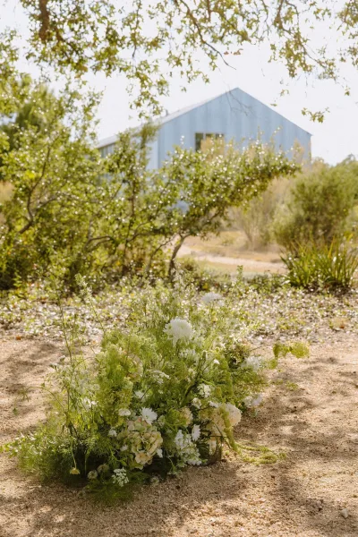 Ground floral arrangement as a freestanding floral installation of white florals along a gravel path, framed by trees and a barn.